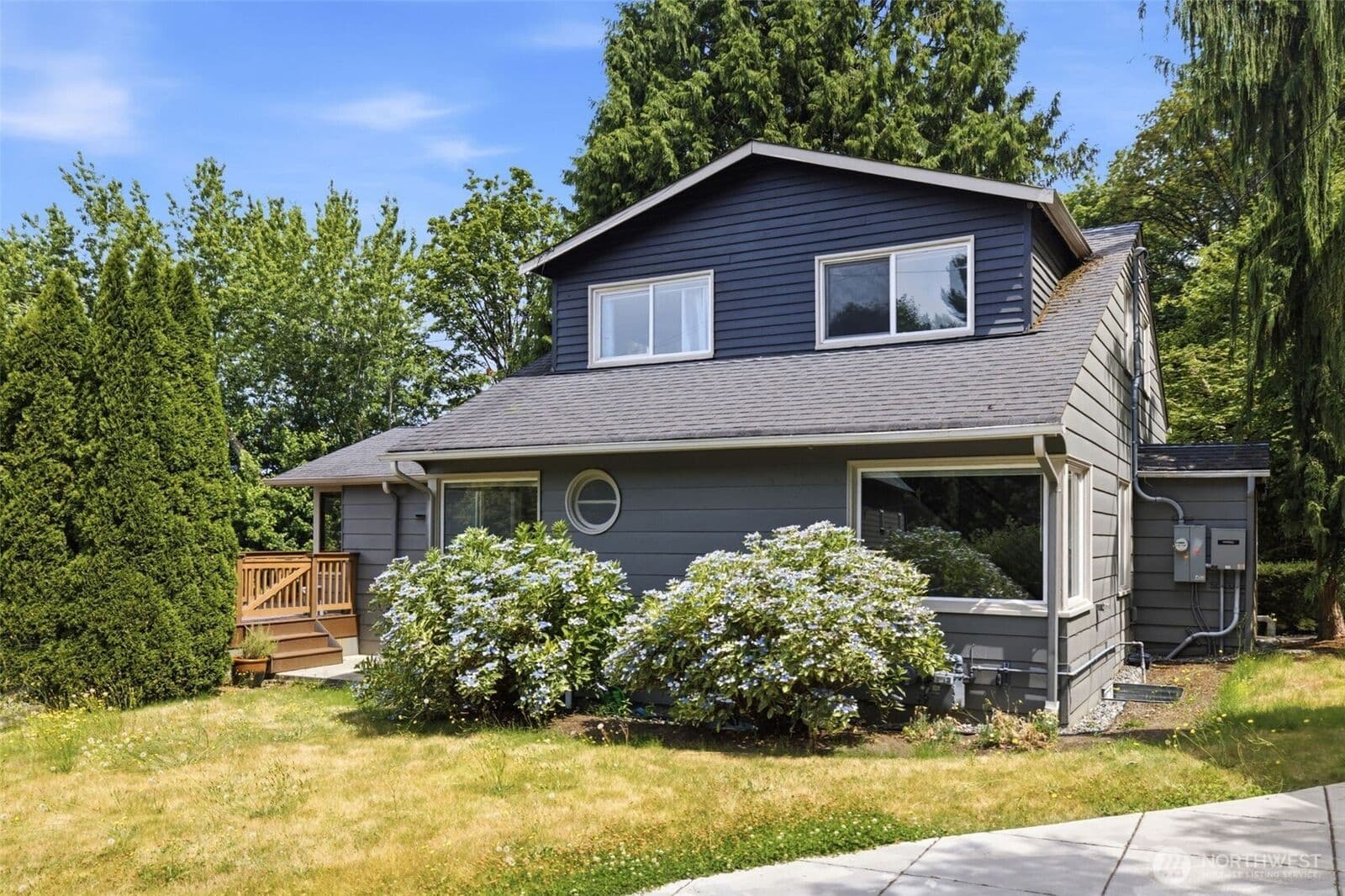 Front elevation of the gray two-story house with white-trimmed windows, a round porthole window, blue hydrangeas along the foundation, and a wooden deck at the left.