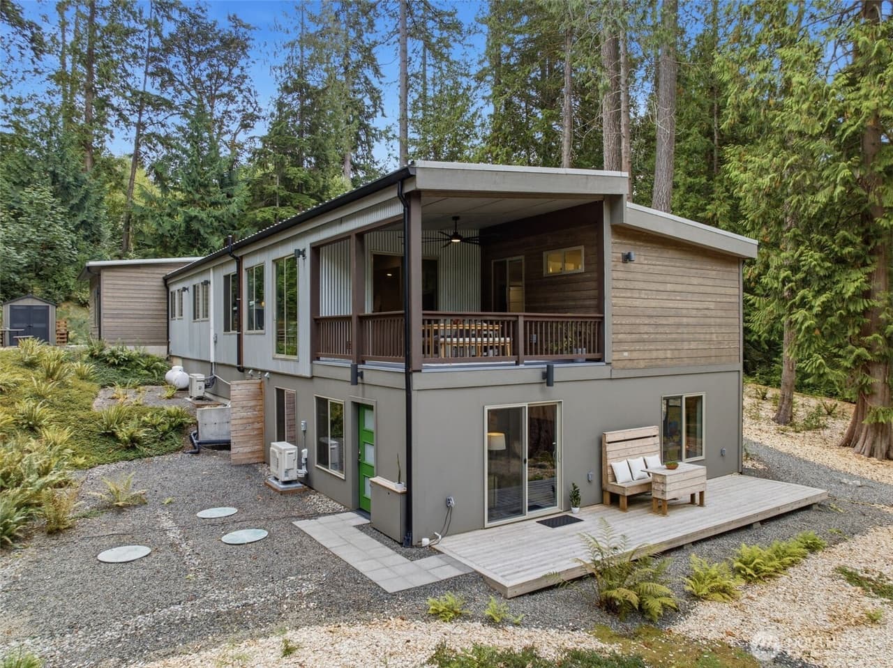 Rear of the two-level modern house in the forest: a covered balcony on the upper level, and the lower level with its own sliding glass door, a separate entry door and a private patio.
